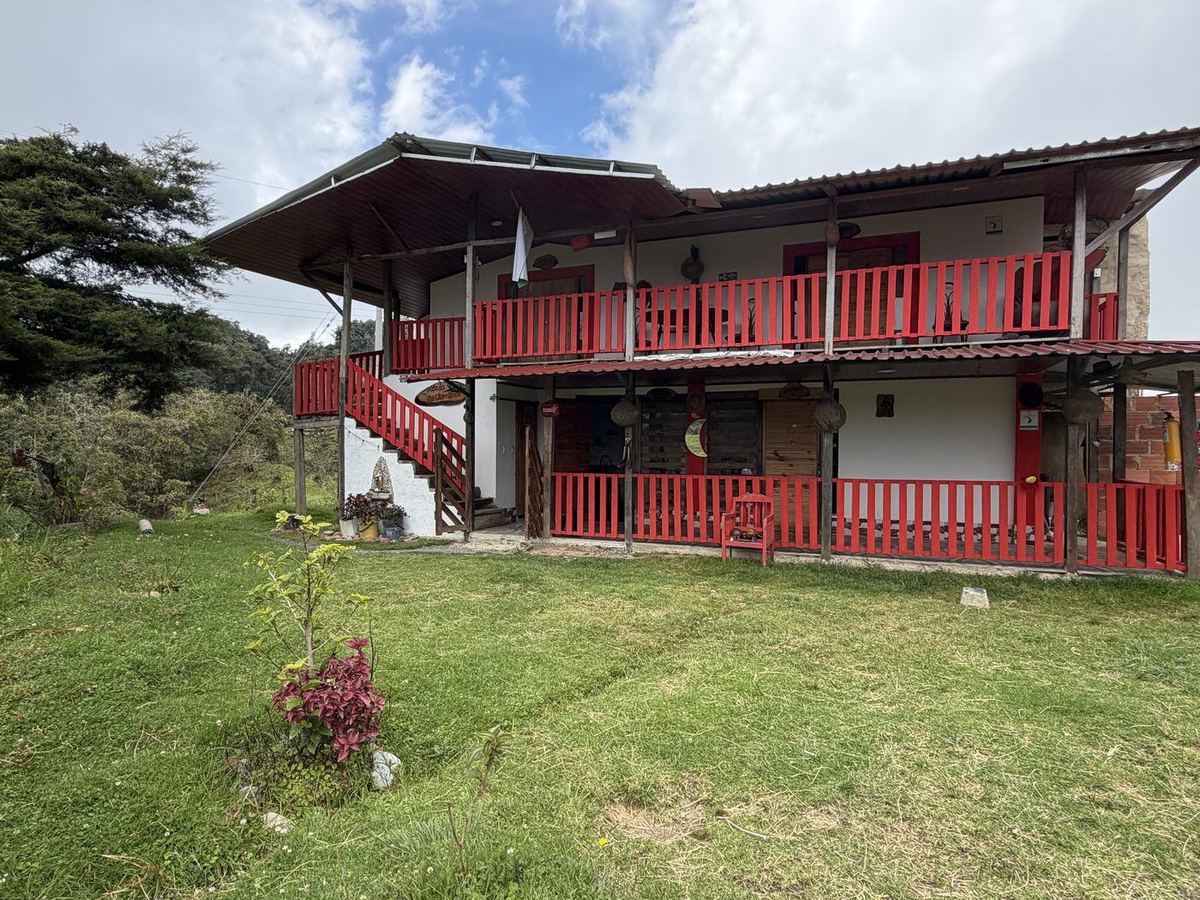 Fachada de dos plantas de la finca Los Clavellinos con corredores de baranda roja, jardín verde y montañas de El Peñón al fondo