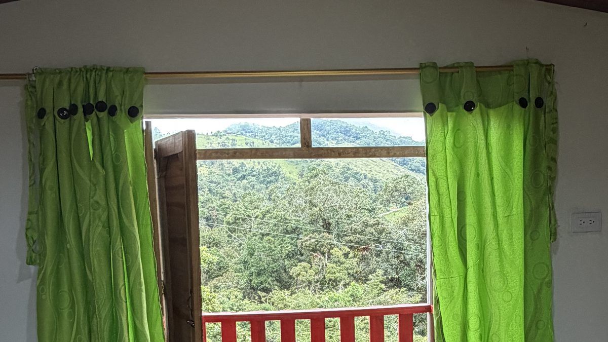 Vista desde el interior de una habitación hacia el balcón de baranda roja y las montañas boscosas de El Peñón, Santander